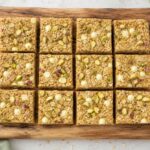 Pistachio white chocolate oat bars cut into squares on a wooden board, showing chewy texture and white chocolate chips.