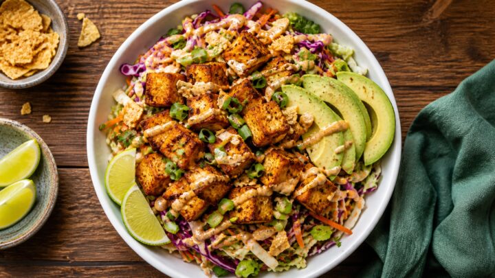 Crispy tofu crunch wrap cabbage slaw bowls with sliced avocado and spicy sauce on a white plate.