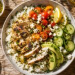 A bowl of olive oil citrus marinated chicken over rice with fresh tomatoes, cucumber, avocado, and crumbled feta cheese.