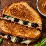 A blackberry basil grilled cheese sandwich with hot honey drizzle on a wooden board, cut in half, showing melted cheese and blackberries.