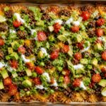 Crispy rice taco salad tray on a baking sheet with seasoned ground beef, melted cheese, lettuce, tomatoes, avocado, black beans, and sour cream.