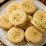 Stack of yuzu pistachio shortbread sandwich cookies with green pistachio pieces and creamy filling, on a rustic wooden board