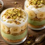 Pistachio banana pudding crunch cups in a clear glass cup showing layers of graham cracker crust, green pudding, and banana slices, topped with whipped cream and chopped pistachios.