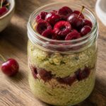 Cherry matcha overnight weetabix in a mason jar with fresh cherries and a wooden spoon.