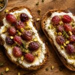 Two slices of pistachio ricotta toast topped with roasted red grapes and chopped pistachios on a wooden board