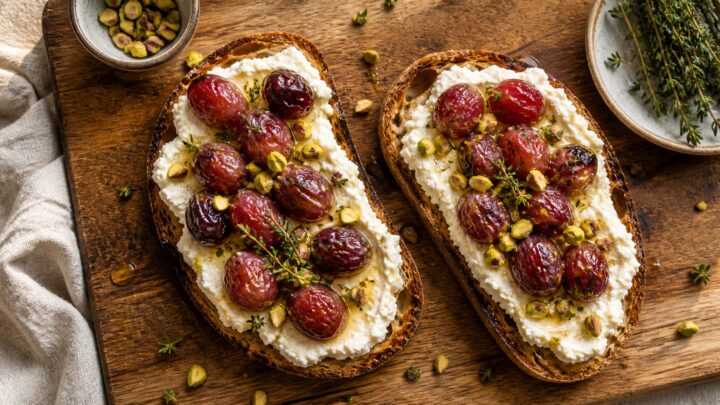 Two slices of pistachio ricotta toast topped with roasted red grapes and chopped pistachios on a wooden board