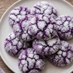 A stack of purple ube coconut crinkle cookies on a white plate, showing their crinkly powdered sugar coating.