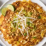 Crispy cabbage pad thai omelet on a plate with lime wedges, topped with peanuts, green onions, and bean sprouts