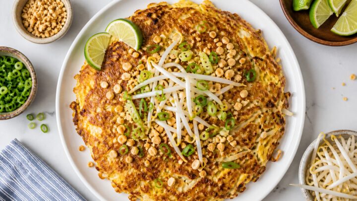 Crispy cabbage pad thai omelet on a plate with lime wedges, topped with peanuts, green onions, and bean sprouts
