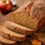 A slice of olive oil apple cider donut loaf on a rustic wooden board with cinnamon sugar coating, surrounded by fall apples and leaves