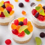 Close up of gummy fruit salad cheesecake cups in clear cups showing graham cracker crust, creamy white filling, and colorful gummy fruit candies on top