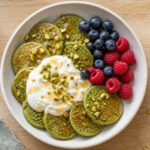 A bowl of small matcha green pancakes topped with Greek yogurt, chopped pistachios, and fresh berries on a wooden table