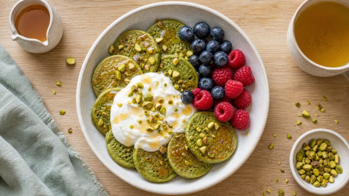 A bowl of small matcha green pancakes topped with Greek yogurt, chopped pistachios, and fresh berries on a wooden table