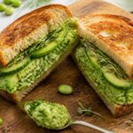 Smashed edamame green goddess sandwich on toasted bread with avocado, cucumber, and sprouts on a wooden cutting board.