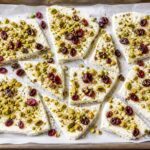 Pistachio labneh breakfast bark on a parchment-lined baking sheet, broken into pieces, with visible pistachios and dried cranberries.