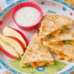 Cheesy veggie toddler lunch quesadilla cut into triangles, on a colorful plate with apple slices on the side.
