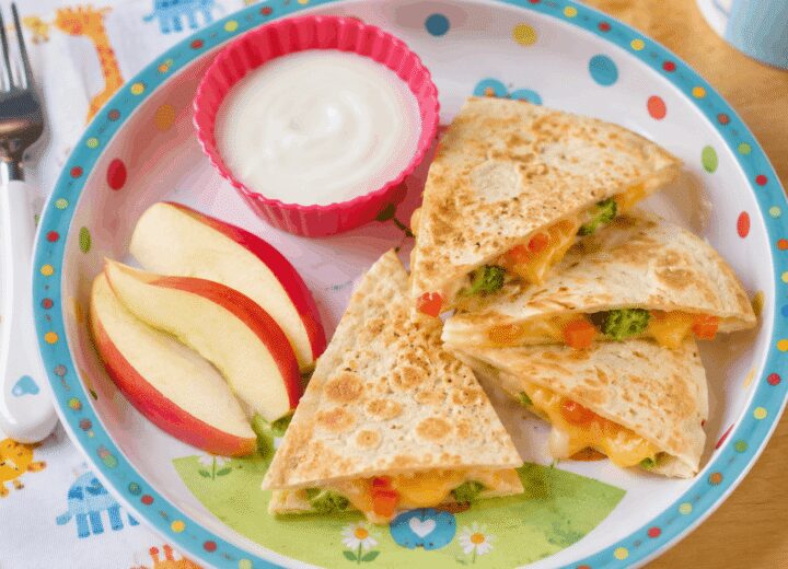 Cheesy veggie toddler lunch quesadilla cut into triangles, on a colorful plate with apple slices on the side.