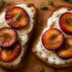 Two slices of roasted plum goat cheese protein toast on a wooden board, with roasted plum halves and fresh thyme garnish.