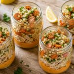 Mason jar layered with roasted carrots, hummus, chicken, and pearl couscous on a wooden table with fresh parsley garnish