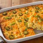 A golden brown cottage cheese jalapeno cornbread breakfast bake in a baking dish, topped with melted cheddar cheese and green onions, served on a rustic wooden table.