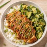 Ginger scallion chicken sliced over white rice in a bowl with smashed cucumber salad on top, garnished with scallions and sesame seeds.