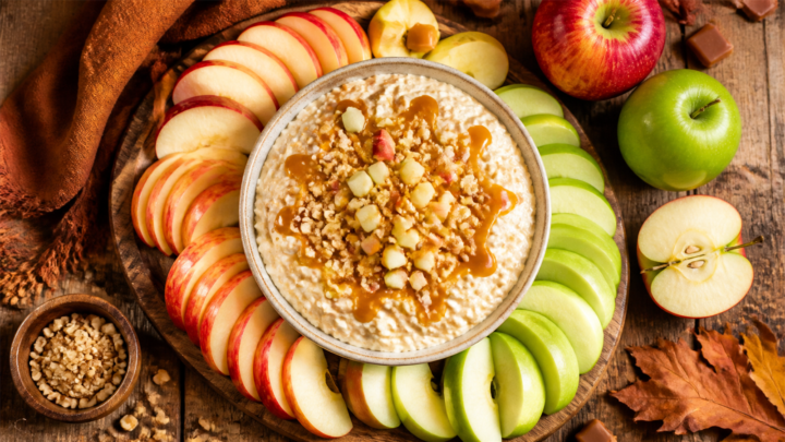 A bowl of creamy caramel apple cottage cheese dip surrounded by fresh apple slices and sprinkled with chopped peanuts on a wooden board.