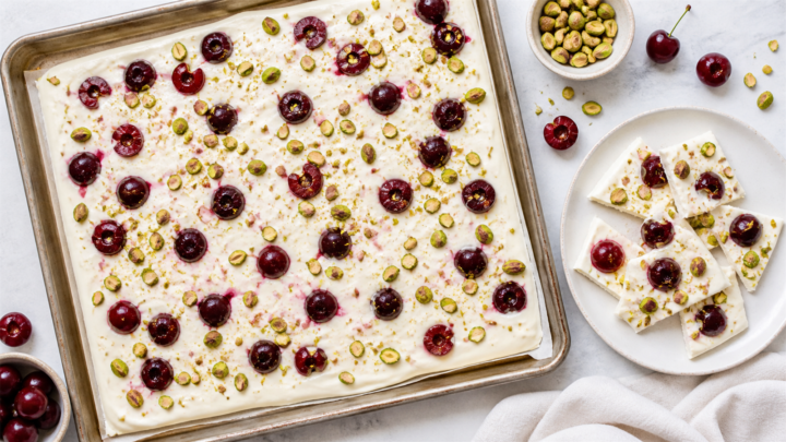 A baking sheet of frozen yogurt bark topped with fresh cherries and chopped pistachios, broken into pieces on a white plate.