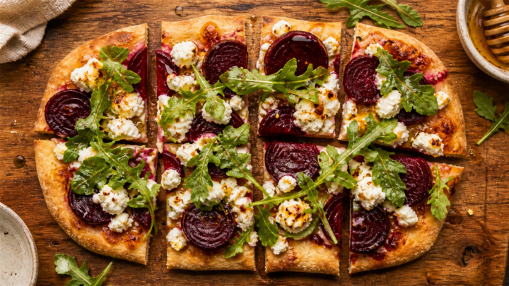 Hot honey goat cheese roasted beet flatbread on a wooden board, sliced, with arugula and hot honey drizzle