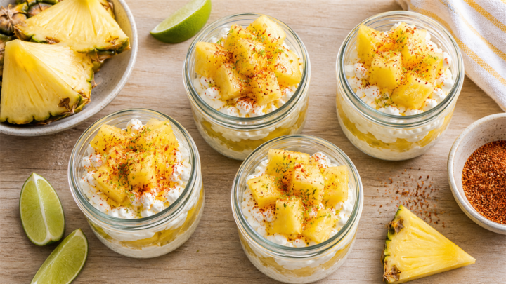 Four individual Pineapple Tajin Cottage Cheese Snack Cups in small jars, topped with Tajin seasoning and lime zest, on a wooden table.
